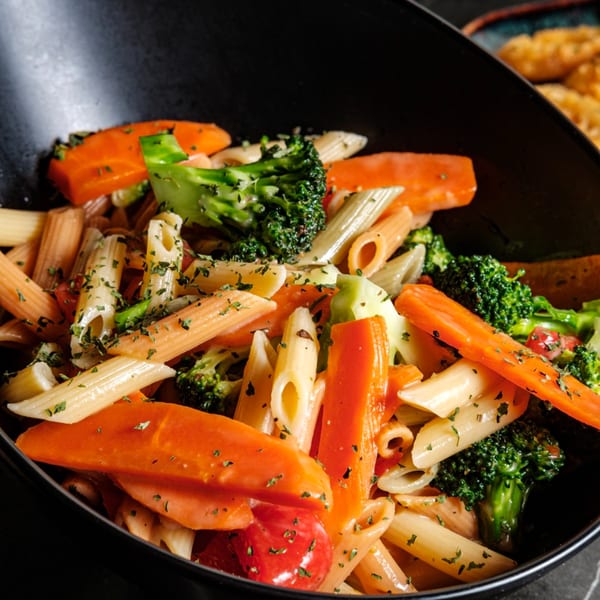 Close-up of penne pasta with broccoli, carrots, and tomatoes, garnished with herbs in a black bowl.