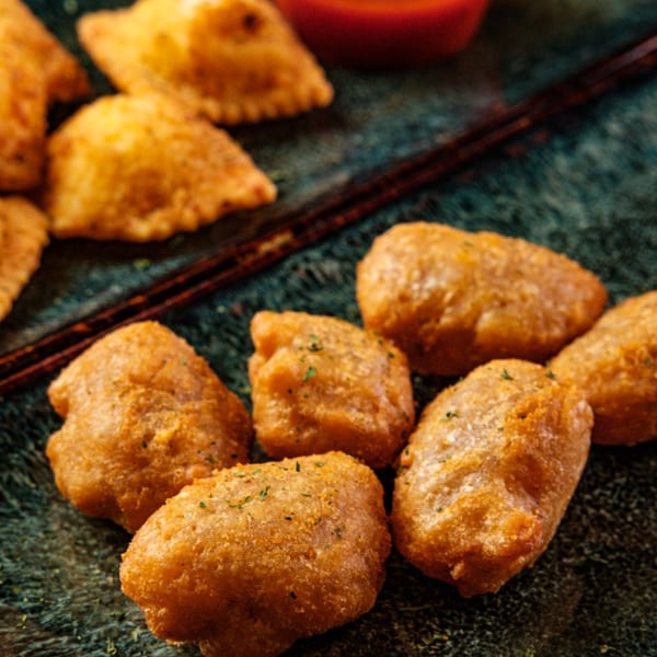 Golden-brown fried pizza bites, some with green herbs, on a dark textured surface with chopsticks and red sauce.